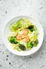 Gourmet shrimp salad with broccoli, cucumber ribbons, and mixed greens, beautifully plated on white dish over stone background