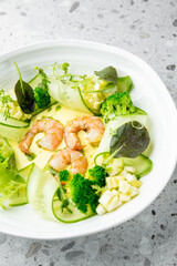 Gourmet shrimp salad with broccoli, cucumber ribbons, and mixed greens, beautifully plated on white dish over stone background