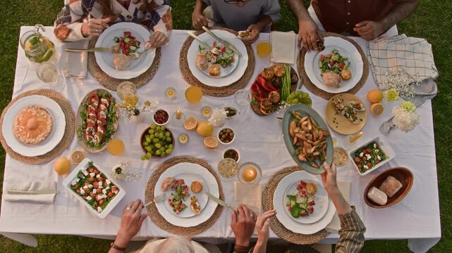 Top view of unrecognizable group of people sitting at dinner table set with white cloth and various dishes enjoying homemade meal during family gathering hands passing dishes over table, copy space