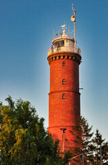 Jarosławiec- Zabytkowa, czerwona, ceglana latarnia morska z tarasem widokowym, otoczona bujną zielenią i pod intensywnie błękitnym niebem o zachodzie słońca. Detale architektoniczne i jaskrawe kolory. © Pol Badger