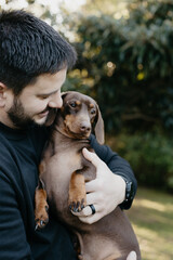 Man with black hair and wearing black holding and cuddling a brown dachshund. Love your pet, dog, companion