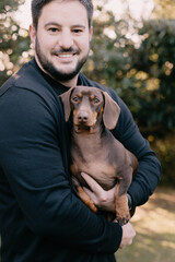 Man with black hair and wearing black holding and cuddling a brown dachshund. Love your pet, dog, companion