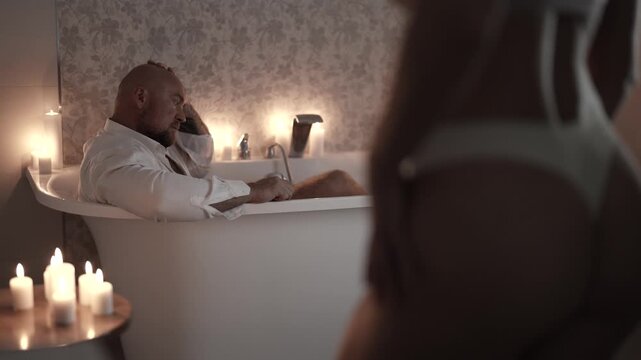 A man with his head bowed in the bathtub, a woman in lingerie stands next to him, candles create an intimate light.