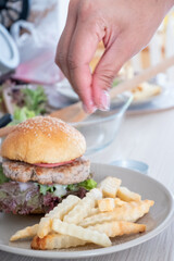 Close-up of a hand sprinkling salt onto french fries next to a homemade burger on a plate, Cooking Mealtime.