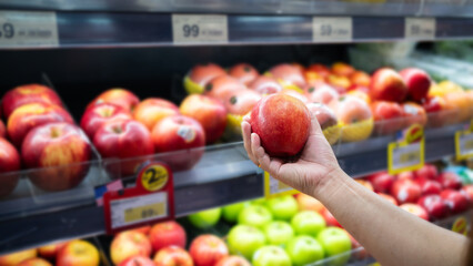 Hand holding fresh red apple in supermarket produce section. Customer selecting ripe fruit from display of colorful apples and green produce for healthy grocery shopping.