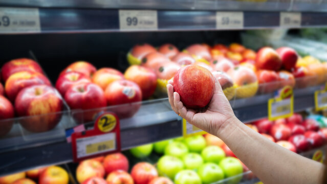 Hand holding fresh red apple in supermarket produce section. Customer selecting ripe fruit from display of colorful apples and green produce for healthy grocery shopping.