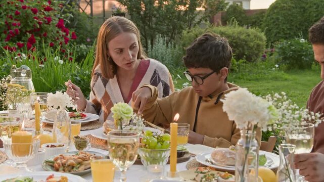 Shot of mother talking to son while sitting at dinner table outdoors during festive gathering in green garden, teaching table manners and talking about food with boy nodding in agreement