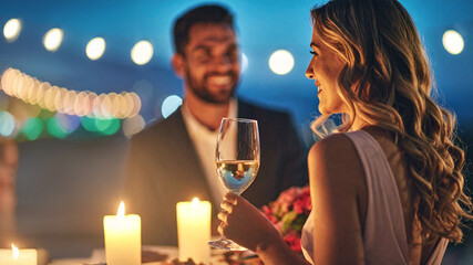 A happy couple on a romantic outdoor dinner date, with the woman holding a glass of white wine. The scene is lit by soft, glowing candles and string lights, creating a warm and intimate atmosphere.