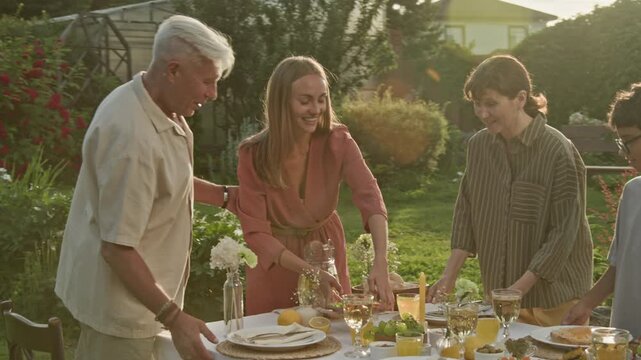 Medium shot of big happy family setting up elegant dinner table outdoors in summer with young woman bringing homemade dishes and smiling