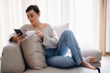 Obraz premium Woman sitting on sofa , drinking tea and using mobile phone