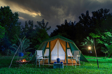 Camping tents on the green grass at night, background with many big trees, vintage cabin tents, antique oil lamps, retro chairs