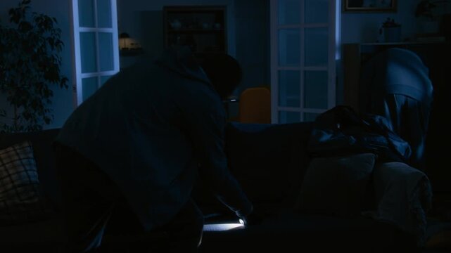 Two unrecognizable accomplices in balaclavas and gloves searching coffee table and checking all bookshelves in living room illuminated by robbers flashlights