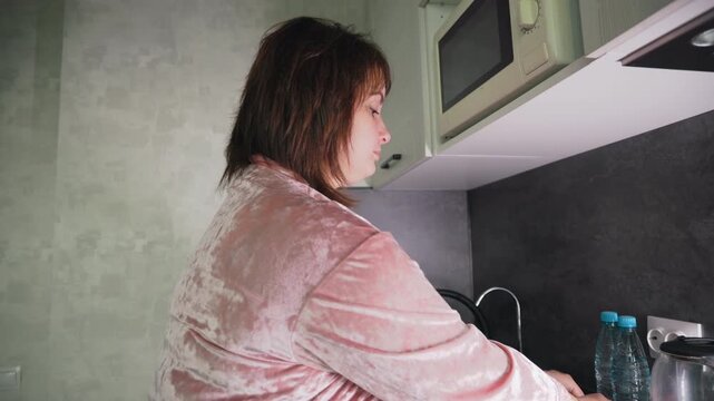 Caregiver in pink robe preparing food at kitchen counter, focused on assembling sandwiches beside bottled water and electric kettle under cabinet with microwave, engaging in early morning routine