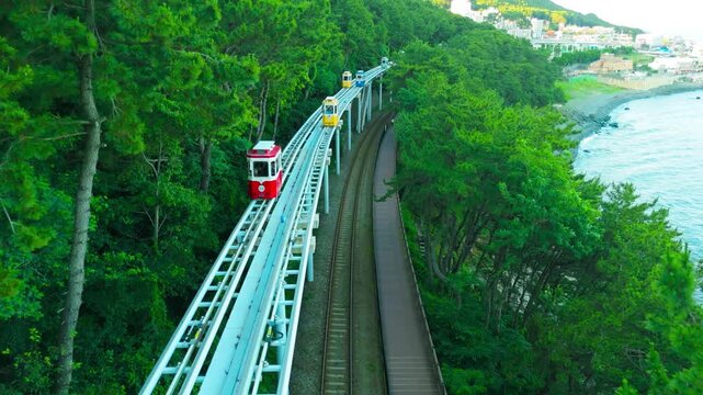 Aerial view of a monorail track with colorful sky capsules winding through a green coastal pine forest, overlooking the ocean and a town in Cheongsapo, Busan, South Korea.