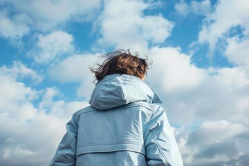Woman admiring cloudy sky with wind blowing her hair, wearing light blue waterproof jacket