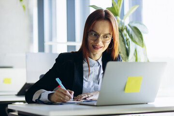 Young business woman sitting at the office using laptop and making notes