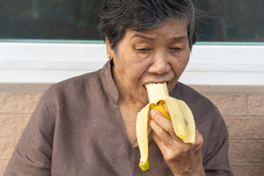 Old Asian woman eating a banana while sitting outdoors, enjoy healthy snack with a joyful expression, promoting wellness, nutrition, simple pleasures in daily life.