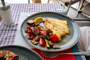 Plate of grilled fish fillet with roasted vegetables and lemon wedge on a restaurant table. Concept of healthy eating, Mediterranean cuisine and seafood