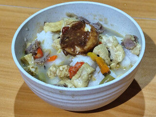 A bowl of vegetable lontong with coconut milk sauce and accompaniments of tofu, beef and Indonesian petis eggs, served on a wooden table.