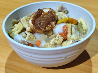A bowl of vegetable lontong with coconut milk sauce and accompaniments of tofu, beef and Indonesian petis eggs, served on a wooden table.