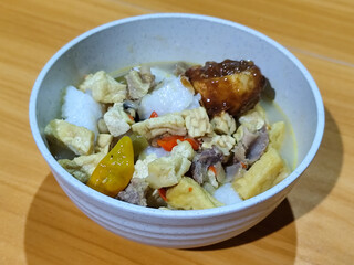 A bowl of vegetable lontong with coconut milk sauce and accompaniments of tofu, beef and Indonesian petis eggs, served on a wooden table.
