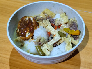 A bowl of vegetable lontong with coconut milk sauce and accompaniments of tofu, beef and Indonesian petis eggs, served on a wooden table.