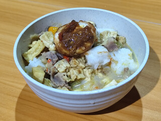 A bowl of vegetable lontong with coconut milk sauce and accompaniments of tofu, beef and Indonesian petis eggs, served on a wooden table.
