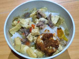 A bowl of vegetable lontong with coconut milk sauce and accompaniments of tofu, beef and Indonesian petis eggs, served on a wooden table.