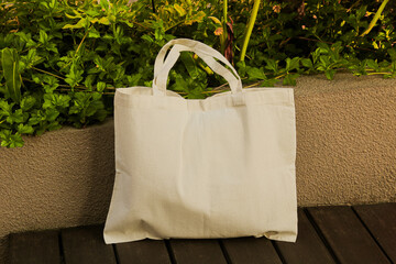 A blank white tote bag is propped against a wall, with green foliage in the background, creating a...