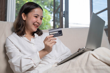 A smiling woman holds a credit card while using a laptop, engaged in online shopping or banking at home.