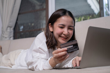 A young woman is lying on a couch, smiling as she uses a laptop and holds a credit card, suggesting online shopping or banking.