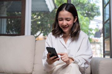 A young woman smiles while using her smartphone on a couch in a bright, comfortable living room, showcasing a casual, relaxed lifestyle.
