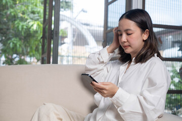 A woman in a white shirt sits on a couch, looking at her smartphone while surrounded by greenery and natural light.