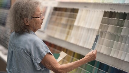 Elderly woman browses paint samples at a hardware store, contemplating various colors for her next...