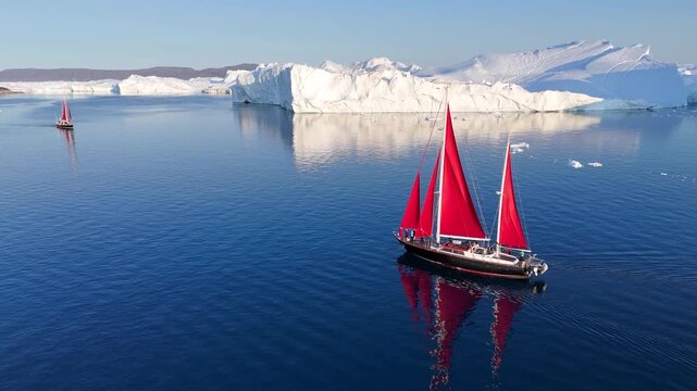 Little red sailboat cruising among floating icebergs in Disko Bay glacier during midnight sun season of polar summer. Ilulissat, Greenland. Global warming and melting glaciers.