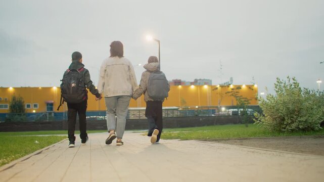 mum holding hands with two children walking down urban park path at dusk capturing family bonding support during evening stroll in green public space near residential area