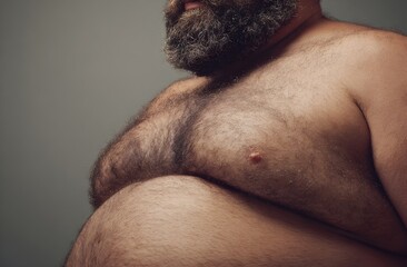 Naklejka premium Bearded overweight man with a hairy chest and large belly, standing against a plain gray background, representing the concept of excess weight and body diversity.
