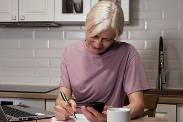 Focused Senior Woman Engaged in Multi-tasking with Phone and Notepad at Kitchen Table, Balancing...