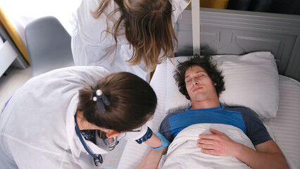 Two nurses bent over a patient lying on a hospital bed. Two female interns and a doctor are going...