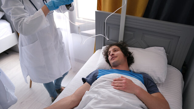 A man is lying in bed, and a doctor is standing next to him. The guy is smiling and in a good mood. A nurse puts an IV drip on a patient in a hospital bed