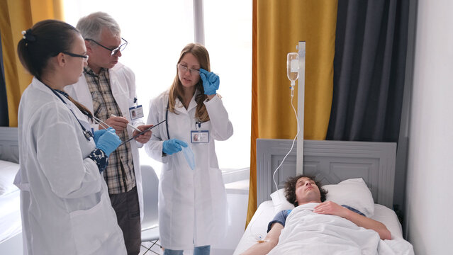 A young female intern in glasses and a white lab coat informs the therapist about the patient's treatment. A man is lying on a hospital bed under an IV drip