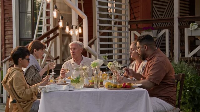 Wide angle scene of modern big family enjoying dinner outdoors and senior man speaking while hosting party with house in background