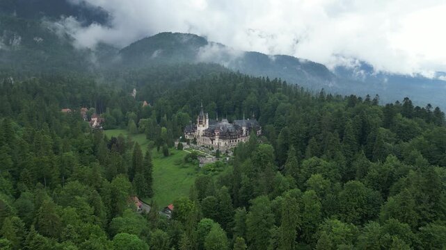 Aerial view of peles castle surrounded by lush greenery and majestic mountains under a cloudy sky, sinaia, prahova, romania.