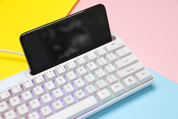 Modern white keyboard with RGB lighting and smartphone on color background, closeup