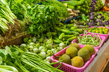Assorted fresh vegetables and tropical fruits displayed in baskets for sale at market stall. Display of variety of leafy greens, cucumbers, eggplants, long beans and green santol on Phuket, Thailand