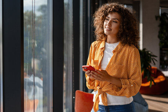 Startup founder holding smartphone looking out window enjoying sunlight in modern office