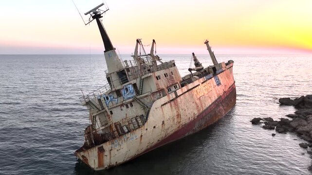Location data omitted, Cyprus - 07 May 2025: Aerial view of an old shipwreck listing in the sea near a rocky coastline, bathed in the warm glow of the rising sun.