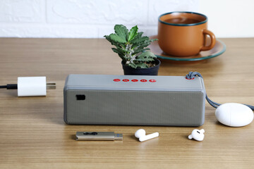A Bluetooth speaker sits between a flash drive, wireless headphones, a small potted succulent plant, and a cup of hot brown tea on a wide table.

