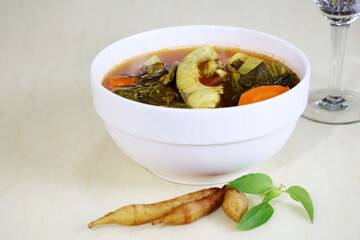 A spicy and sour soup with pieces of fish placed between green and orange vegetables in a white bowl on a cream-colored table.

