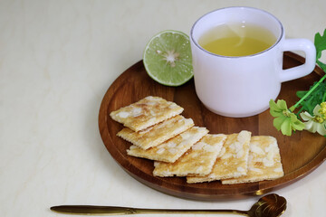 Crackers are arranged on a tray next to cups of hot tea, and a lemon halve sits on a cream-colored table.

